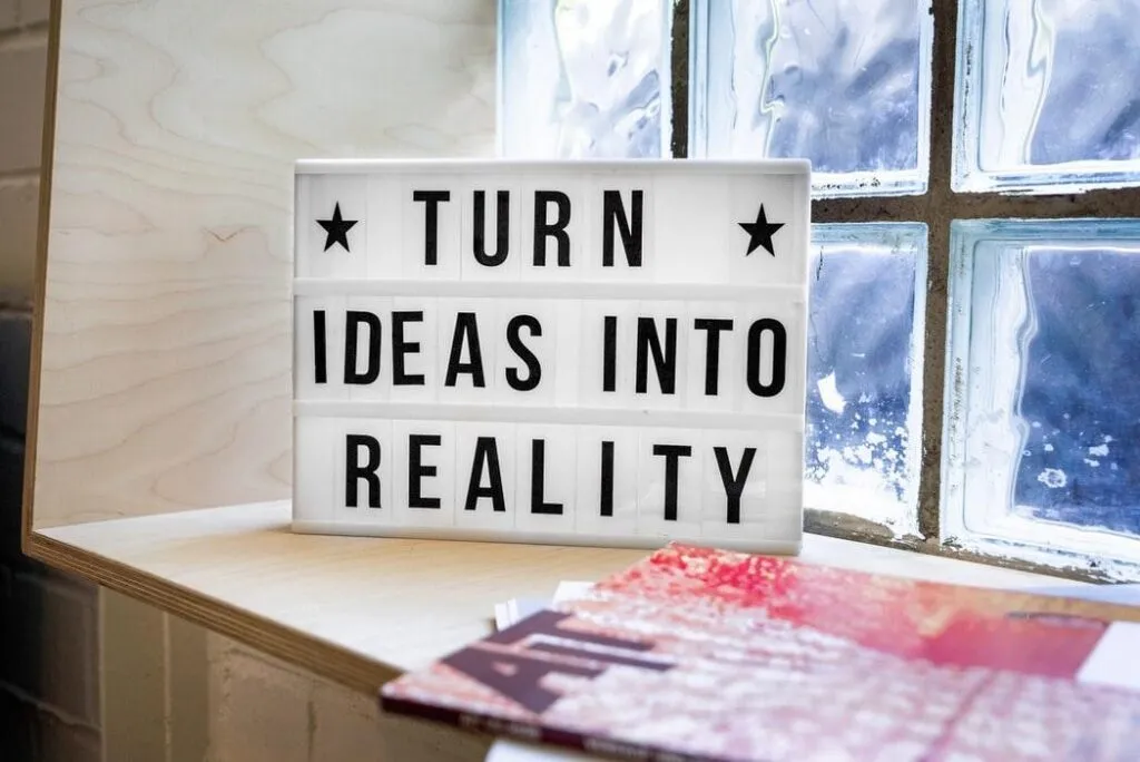 „Leuchtbox mit der Aufschrift ‚TURN IDEAS INTO REALITY‘ steht auf einem Holzregal neben Magazinen vor einem Glasbausteinfenster – Symbol für Innovation und Umsetzungskraft.“