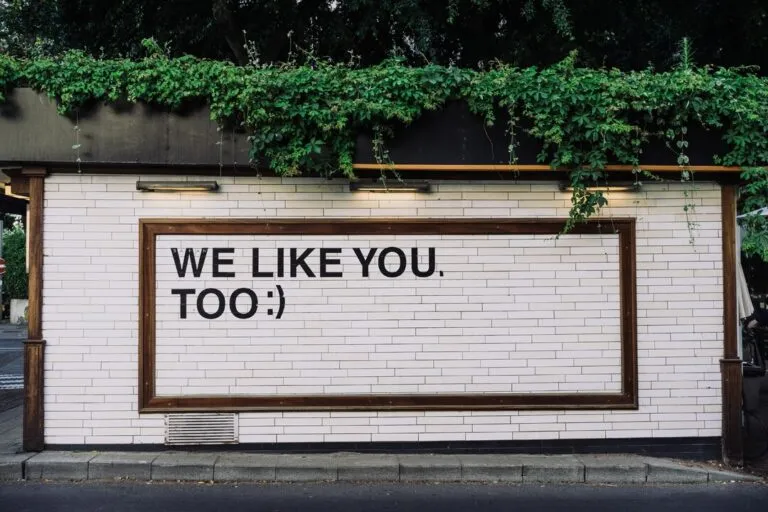 Weiße Wand mit schwarzer Aufschrift ‚WE LIKE YOU TOO :)‘, eingerahmt von Holz und umgeben von grünen Pflanzen – positive Botschaft im urbanen Umfeld