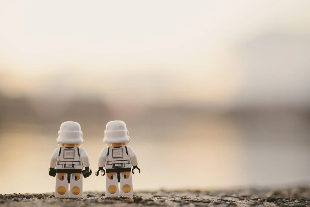 Zwei Lego-Figuren in weißen Anzügen stehen nebeneinander auf einer rauen Oberfläche, mit unscharfem Hintergrund in warmen Farbtönen.