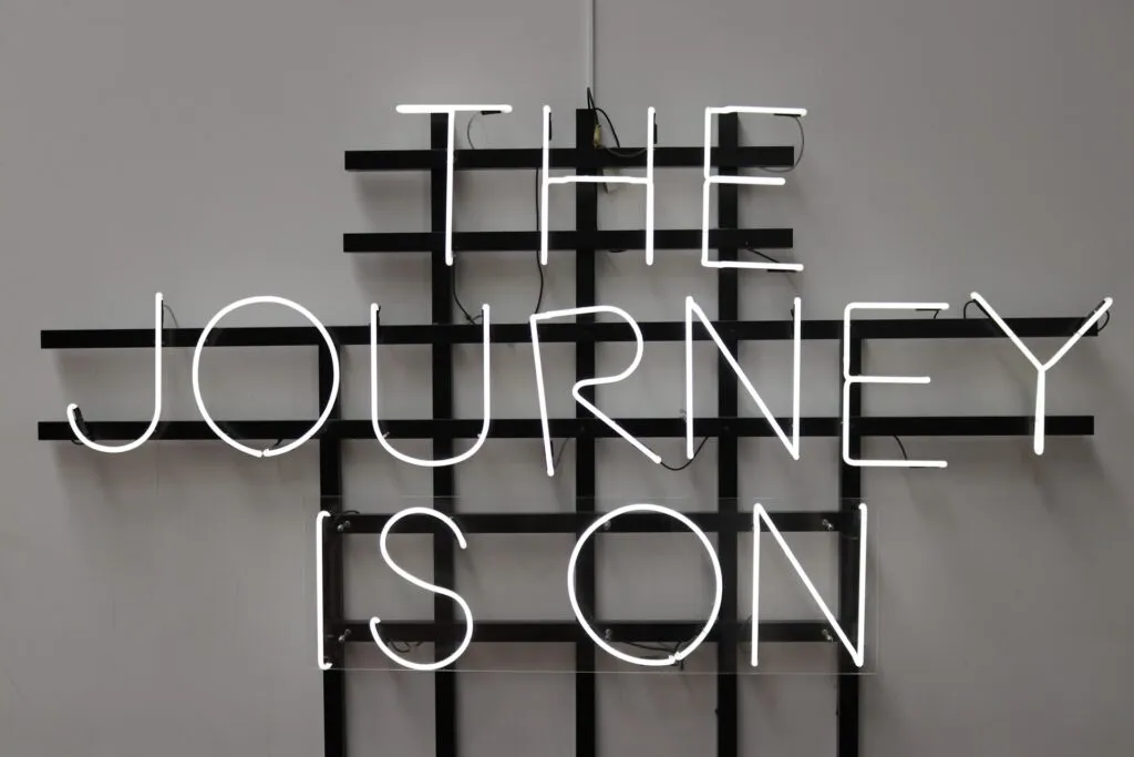 Neon-Schriftzug an einer Wand mit dem Text ‚THE JOURNEY IS ON‘, montiert auf einem schwarzen Gitter.