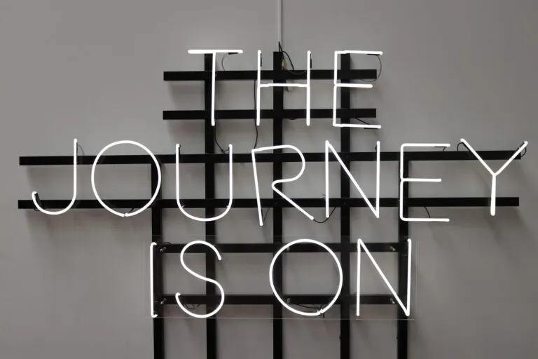 Neon-Schriftzug an einer Wand mit dem Text ‚THE JOURNEY IS ON‘, montiert auf einem schwarzen Gitter.