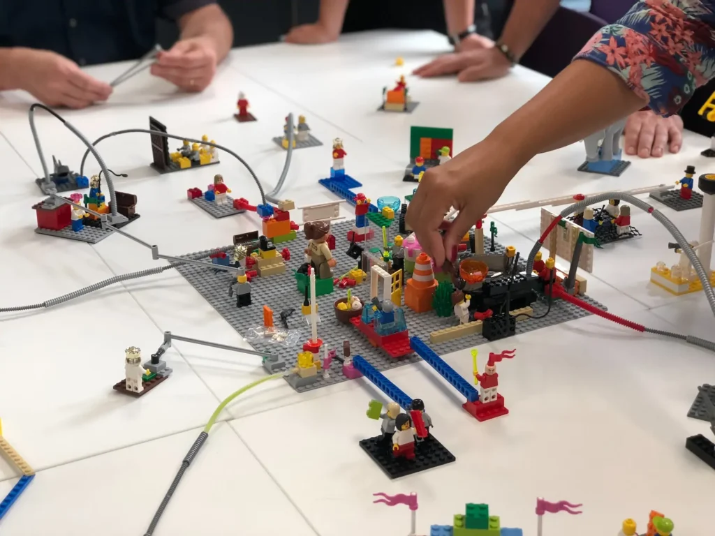 „Personen arbeiten gemeinsam an einem Lego-Modell auf einem Tisch, das aus bunten Bausteinen und Figuren besteht und verschiedene Strukturen darstellt.