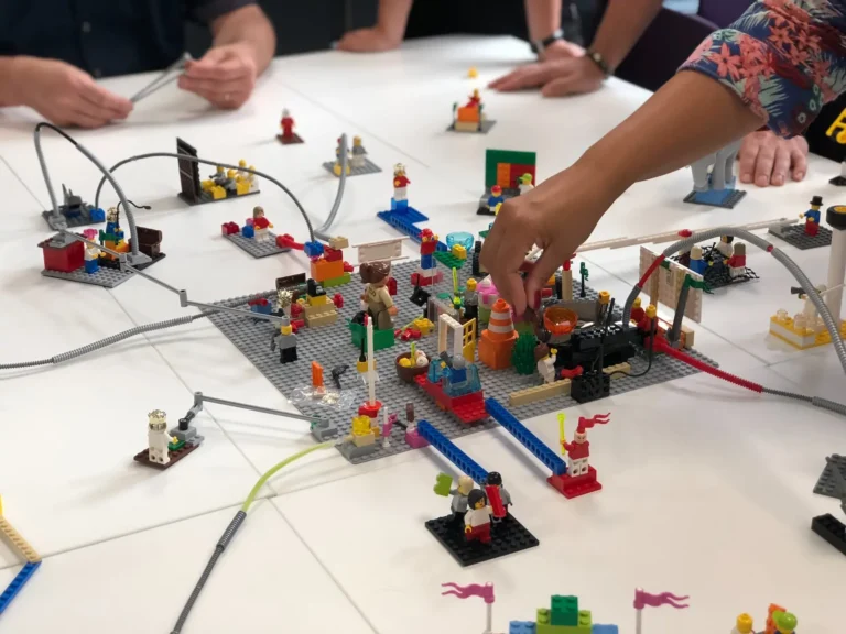 „Personen arbeiten gemeinsam an einem Lego-Modell auf einem Tisch, das aus bunten Bausteinen und Figuren besteht und verschiedene Strukturen darstellt.
