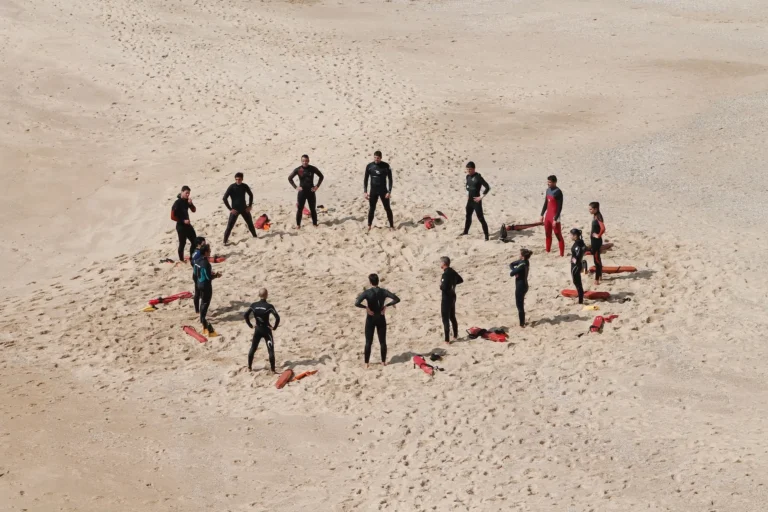 Gruppe von Menschen in Neoprenanzügen steht im Kreis auf einem Sandstrand, umgeben von Rettungsbrettern.