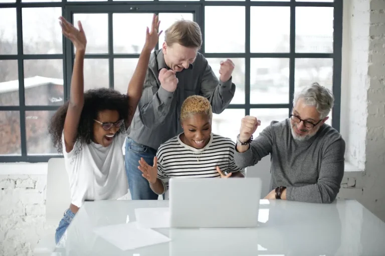 Vier Personen jubeln gemeinsam vor einem Laptop in einem modernen Büro mit großen Fenstern.