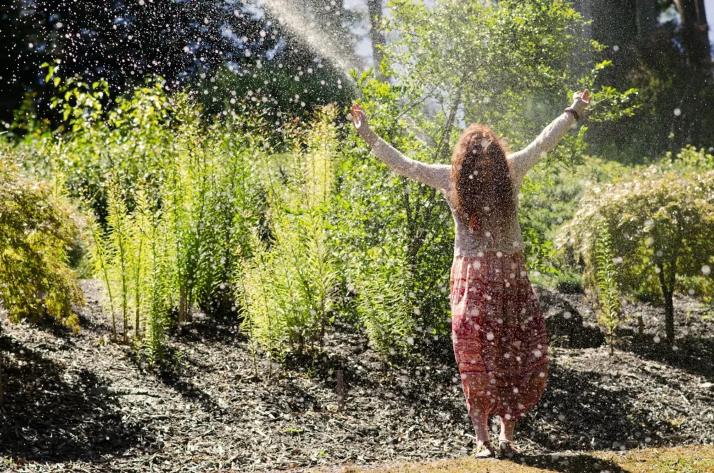 Person steht in einem sonnigen Garten mit ausgebreiteten Armen, während Wassertröpfchen in der Luft glitzern – Symbol für Befreiung und Klarheit