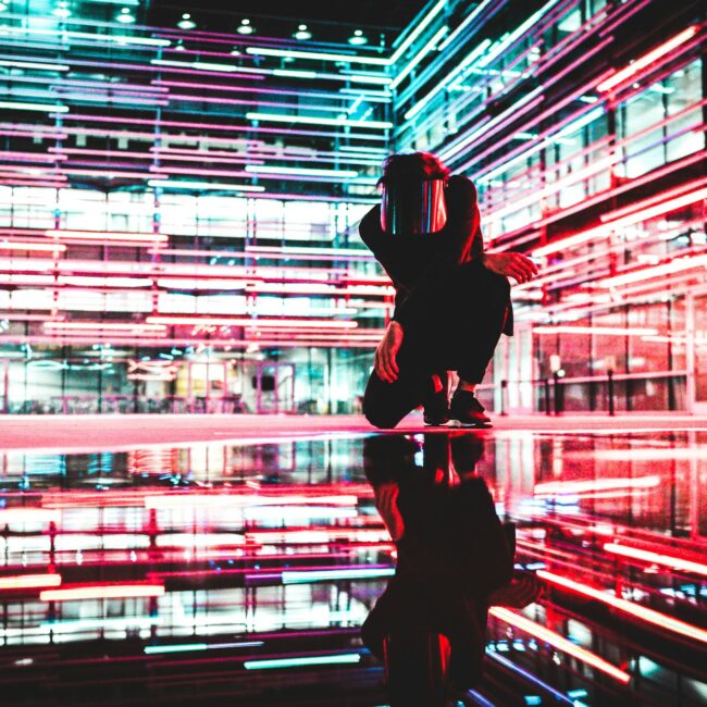 Eine Person in futuristischer Umgebung mit neonfarbenen Lichtlinien, umgeben von reflektierenden Flächen in einem technisch anmutenden Raum.